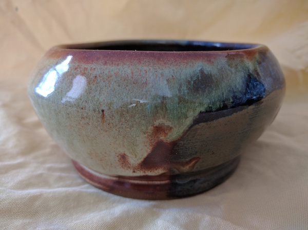 side view of a bowl that is narrow at the bottom, bulges out, and is somewhat narrower at the top, glazed in sea green, rusty brown, and blue