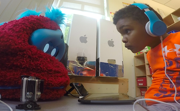 a boy sits at a table with a fluffy robot on it and leans in to peer at the robot's face, while the robot looks down at a tablet