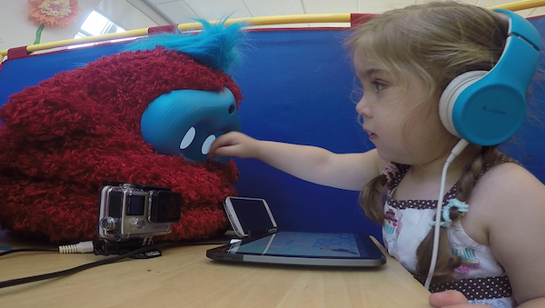 a girl reaches her hand toward the face of a fluffy red robot, which sits on the table in front of her