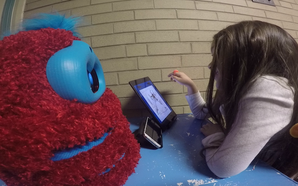 child leans over tablet showing a storybook, in front of a fluffy robot who is listening