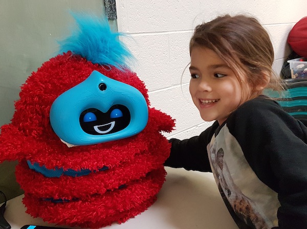 A girl grins at a red and blue fluffy robot and puts her arm around it