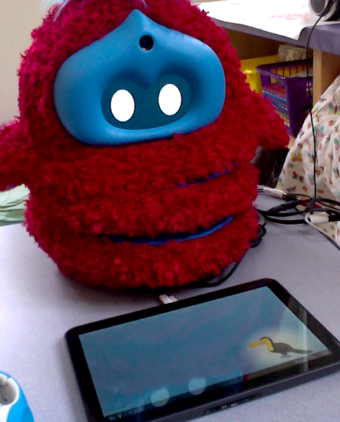 A fluffy red robot sits behind a tablet, which is laying on a table
