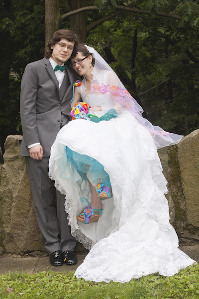 Randy in a tux and Jacqueline in a white wedding dress with colorful accents, in front of a stone wall at a park