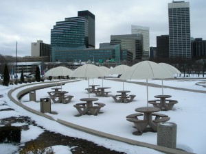 Winter picnic in Cleveland a flock of round picnic tables, cream-colored umbrellas shading benches of snow, with the buildings of Cleveland rising in the background