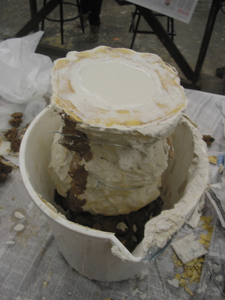 mold tied together, upside-down in a bucket, wet plaster visible in the opening at the neck