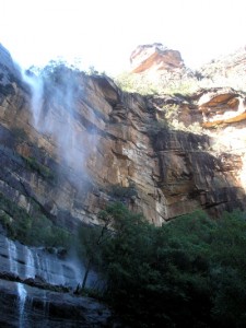 waterfall _spray from a waterfall, red layered rocks above in the sunlight and green bushy trees in the shadows below_