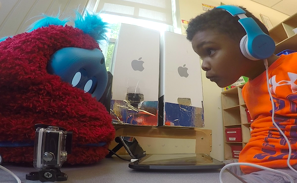 A child plays with Tega a boy sits at a table with a fluffy robot on it and leans in to peer at the robot's face, while the robot looks down at a tablet