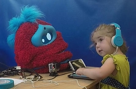 A child mimics with Tega a girl mimics the head tilt and expression shown by a fluffy robot