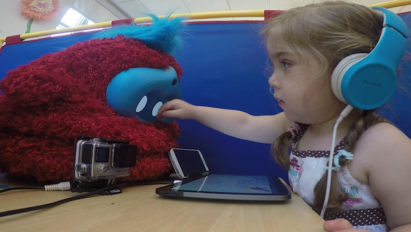Child playing with Tega a girl reaches her hand toward the face of a fluffy red robot, which sits on the table in front of her