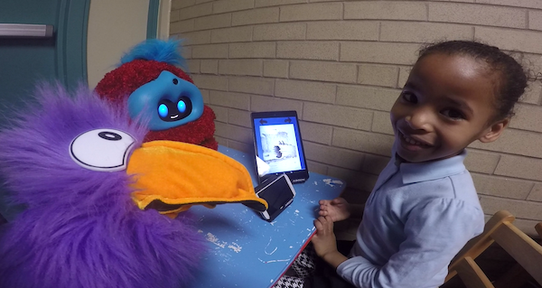 Child plays with Tega robot and puppet child smiling and looking up, beside fluffy robot and fluffy toucan puppet