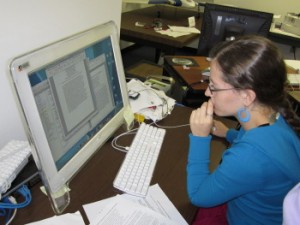 girl in blue sweater sitting at a computer with documents open