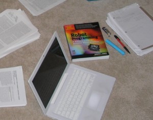 laptop, piles of printed papers, a robot programming text, a highlighter, a flash drive and a pen