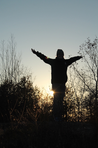 silhouette of a person with arms outstretched on wintery day, in front of bare-limbed trees and a dim sunset sky