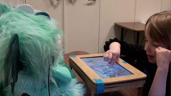 Child playing with storytelling robot Girl moving a picture on a tablet screen, with the tablet inset in a mini table that is between her and a dragon robot