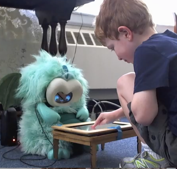 Child playing with storytelling robot Boy touching a screen that is in a mini table that is between him and a dragon robot, the robot is also looking at the table