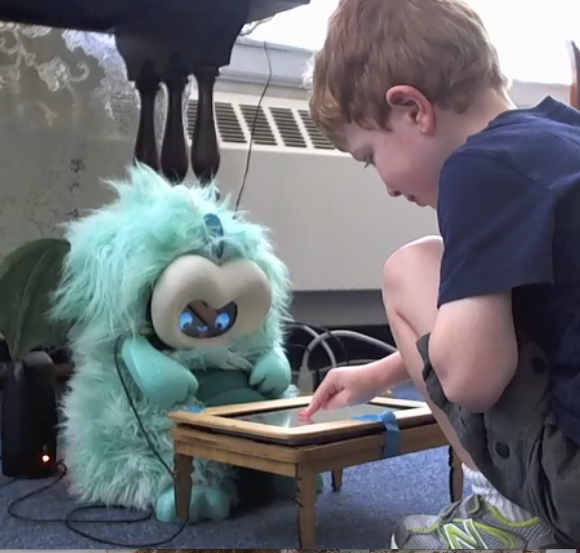 Boy touching a screen that is in a mini table that is between him and a dragon robot, the robot is also looking at the table