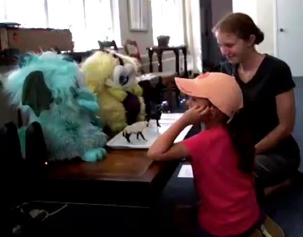 A child and a woman sit in front of a small table, looking at and talking with two fluffy dragon robots that are on the table